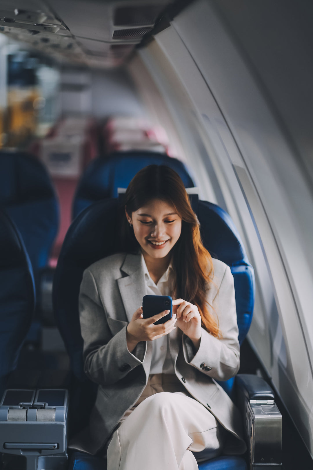 Lächelnde Frau sitzt im Flugzeug und tippt etwas auf ihrem Handy